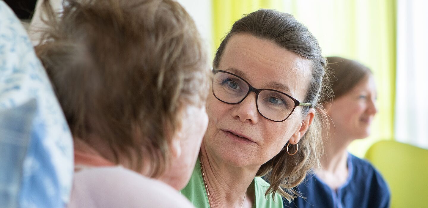 Eine Bewohnerin ist leicht angeschnitten von hinten zu sehen. Eine Pflegefachkraft sitzt neben ihr, wendet sich ihr zu und spricht mit ihr.