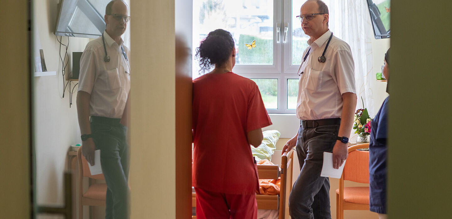 In einem Bewohnerzimmer unterhalten sich der Palliativmediziner und eine Pflegefachkraft während der ärztlichen Visite.