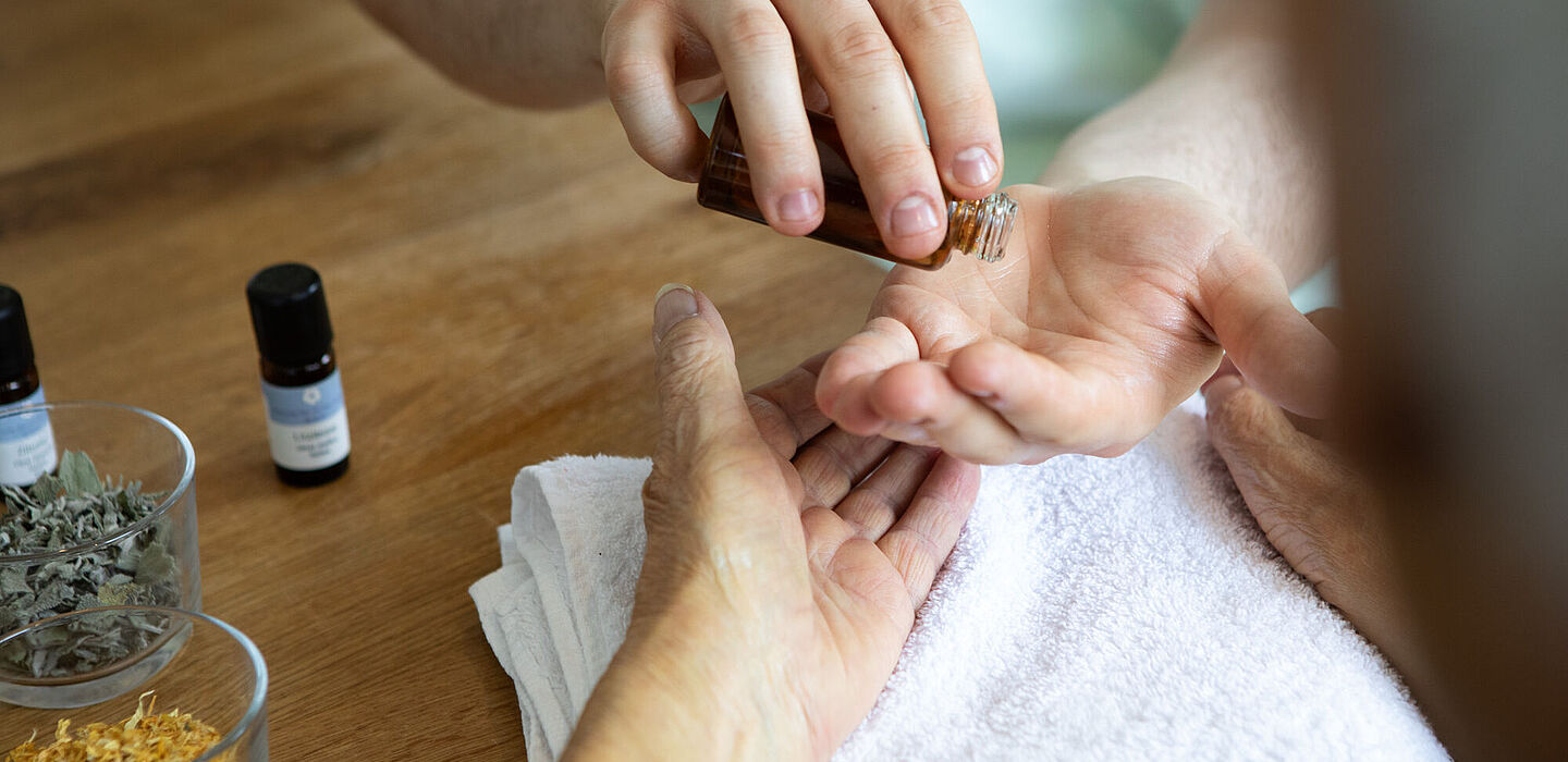 Ein Pfleger gießt Aromaöl für eine Handmassage in seine eigene Hand. Daneben stehen Kräuter und Öl für die weitere Behandlung bereit.