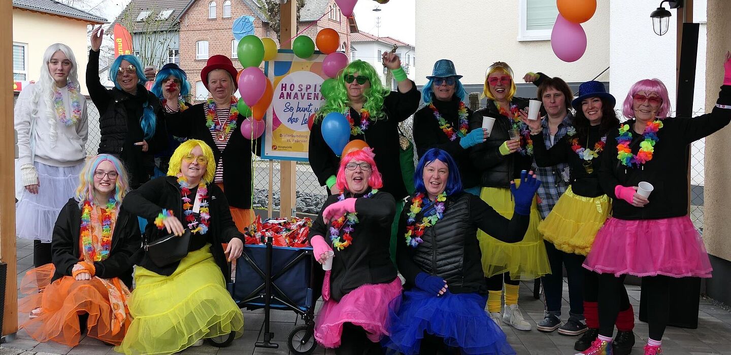 Das Team vom Hospiz ANAVENA 2024 beim Karnevalsumzug. Alle tragen bunte Röcke und bunte Perücken. In der Mitte der Gruppe steht ein Bollerwagen mit dem Schild "Hospiz ANAVENA - Bunt wie das Leben".