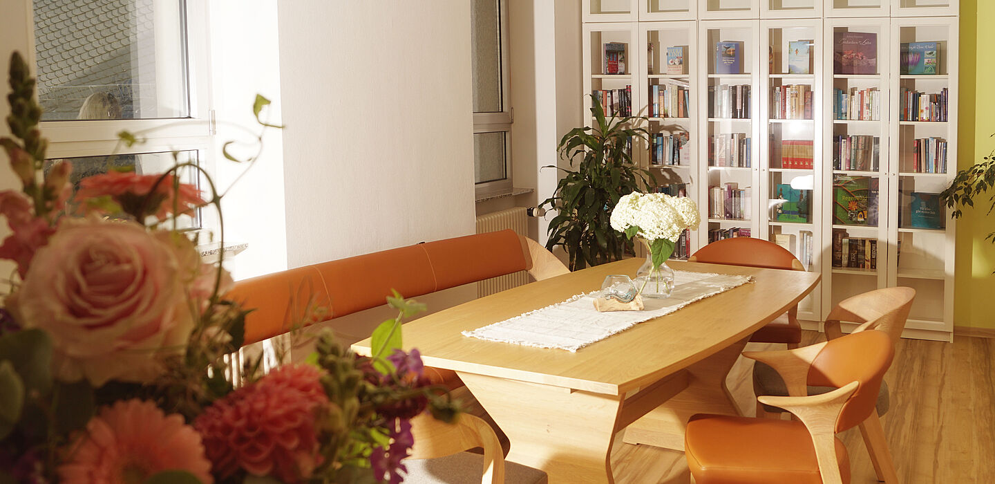 Blick in das Wohnzimmer des Hospizes mit einem großen Holztisch, gemütlichen Stühlen und einer Bücherwand im Hintergrund.