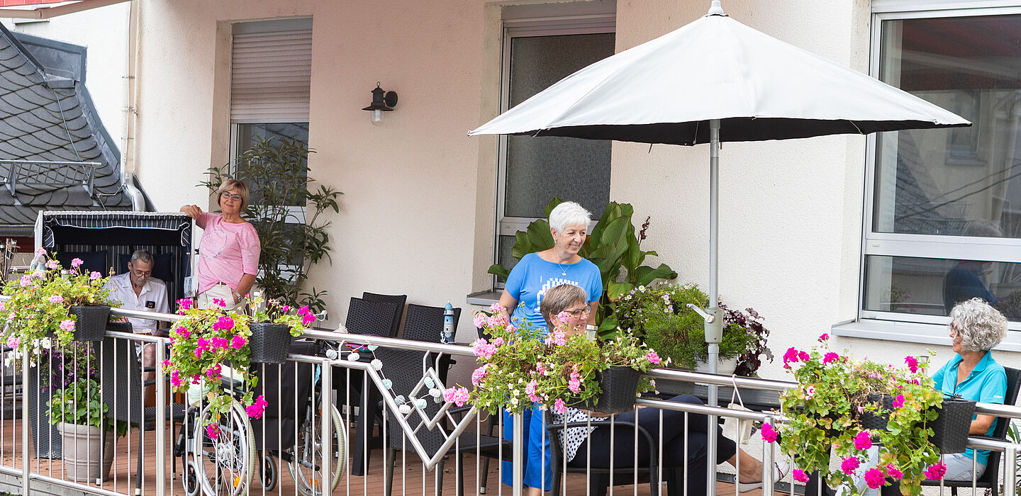 Die Terrasse des Hospizes von außen fotografiert. Man sieht Menschen, einen aufgespannten Sonnenschirm sowie viele blühende Blumen.