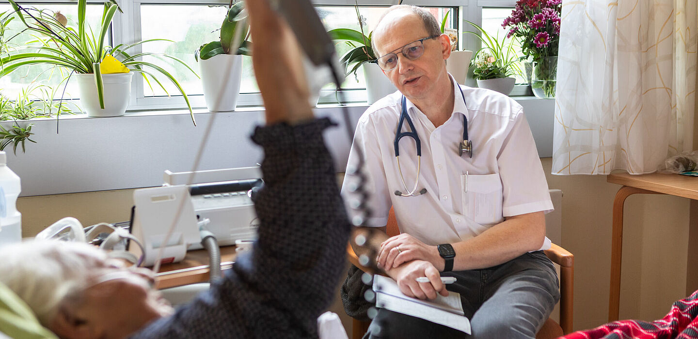 Während der ärztlichen Visite unterhält sich der Palliativmediziner mit einer Bewohnerin. Er sitzt auf einem Stuhl am Fenster, sie liegt im Pflegebett.