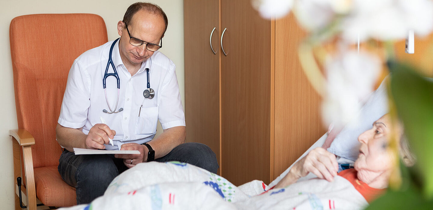 Im Bett liegt eine Bewohnerin. Der Palliativmediziner sitzt daneben auf einem Sessel und notiert etwas auf einem Block, der auf seinem Knie liegt.