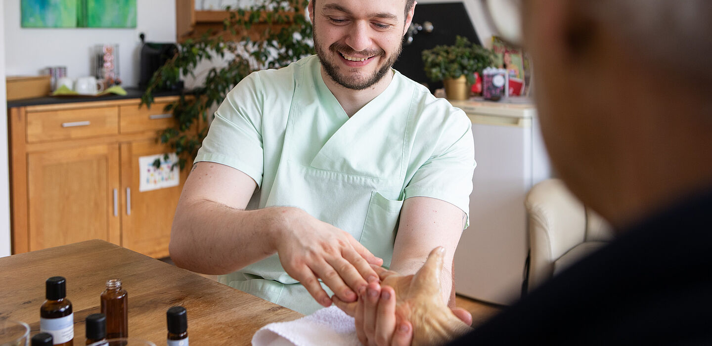 Die Pflegefachkraft bereitet eine Handmassage mit Aromaölen vor. Der Bewohner ist angeschnitten von hinten im Vordergrund zu sehen.