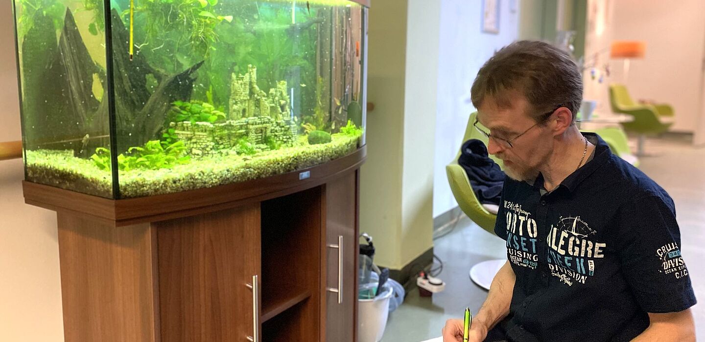 Ein Ehrenamtlicher kümmert sich um das Aquarium im Flur des Hospizes. Er kniet davor und notiert etwas in einem Formular.