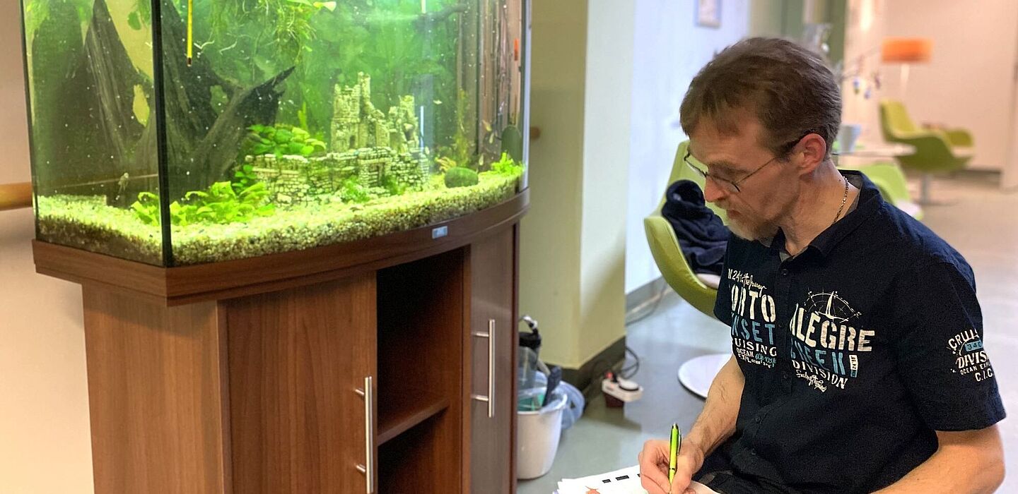 Ein Ehrenamtlicher kümmert sich um das Aquarium im Flur des Hospizes. Er kniet davor und notiert etwas in einem Formular.