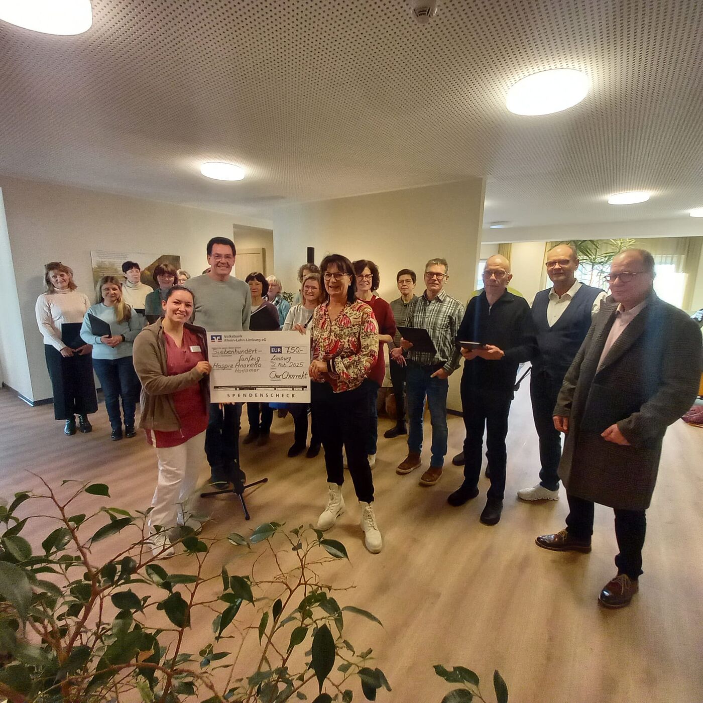 Im Hintergrund stehen die Mitglieder des Chors, im Vordergrund überreichen 2 Vertreter des Chores einen symbolischen Spendenscheck an die stellvertretende Pflegedienstleiterin Sabrina Schleifer.
