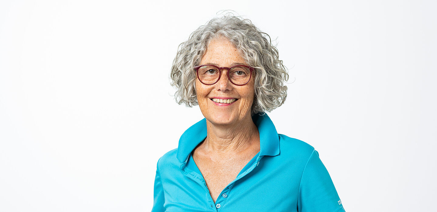 Margit Geiger trägt ein türkisfarbenes Polo-Shirt, eine Brille mit roter Fassung und die grauen Locken auf Kinnlänge offen.