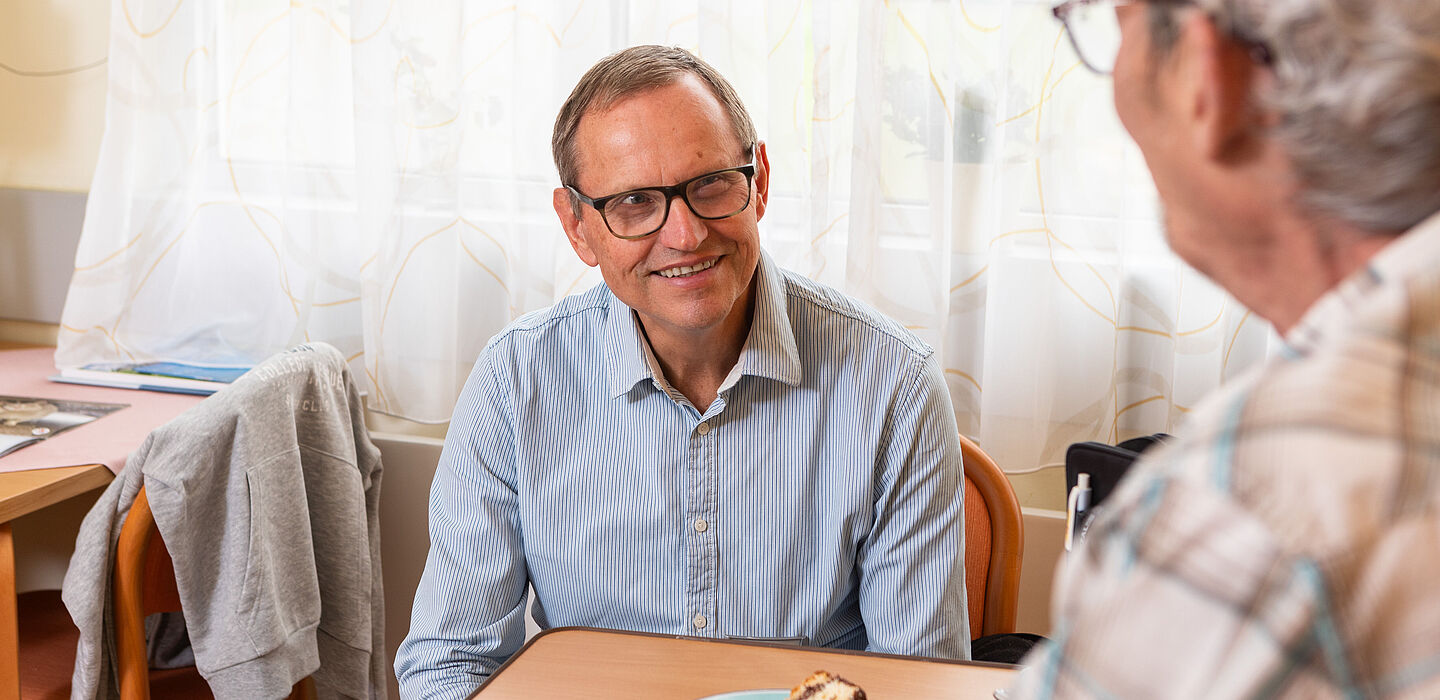 Ein ehrenamtlicher Hospizbegleiter hat einem Bewohner ein Stück Kuchen gebracht und unterhält sich nun mit ihm.