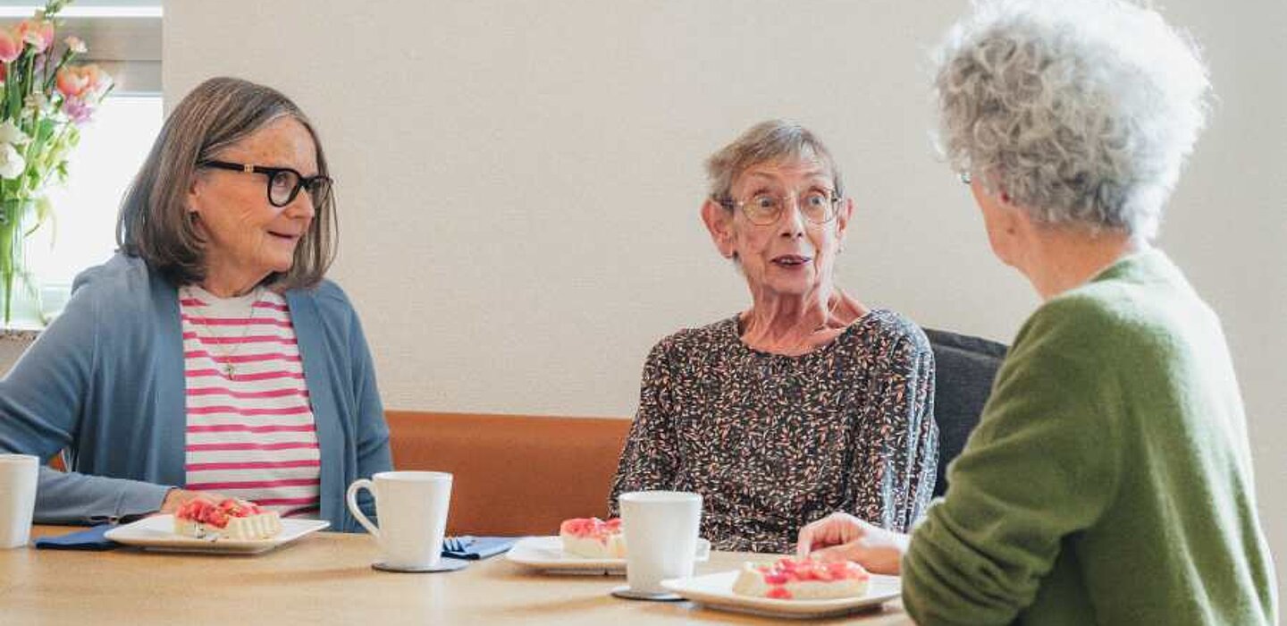 Zwei ehrenamtliche Hospizbegleiterinnen sitzen mit einer Bewohnerin an einem Holztisch, auf dem Kuchenteller und Kaffeetassen stehen. Die drei unterhalten sich.