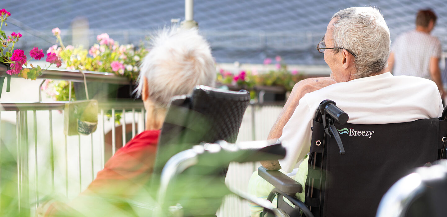 Zwei Bewohner sitzen in Rollstühlen auf der Terrasse und unterhalten sich.