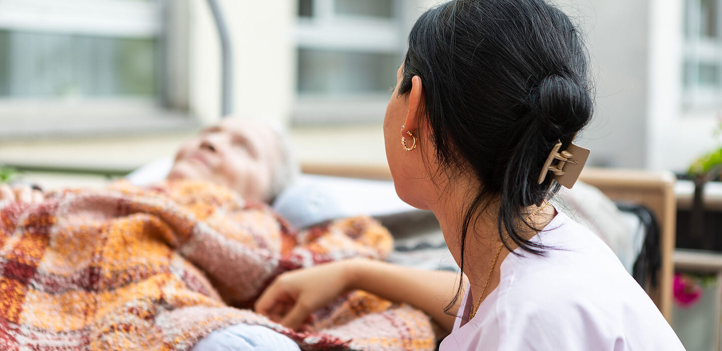 Eine Bewohnerin liegt im Therapiebett, das auf die Terrasse geschoben wurde. Eine Pflegekraft sitzt bei ihr und wendet sich ihr zu.