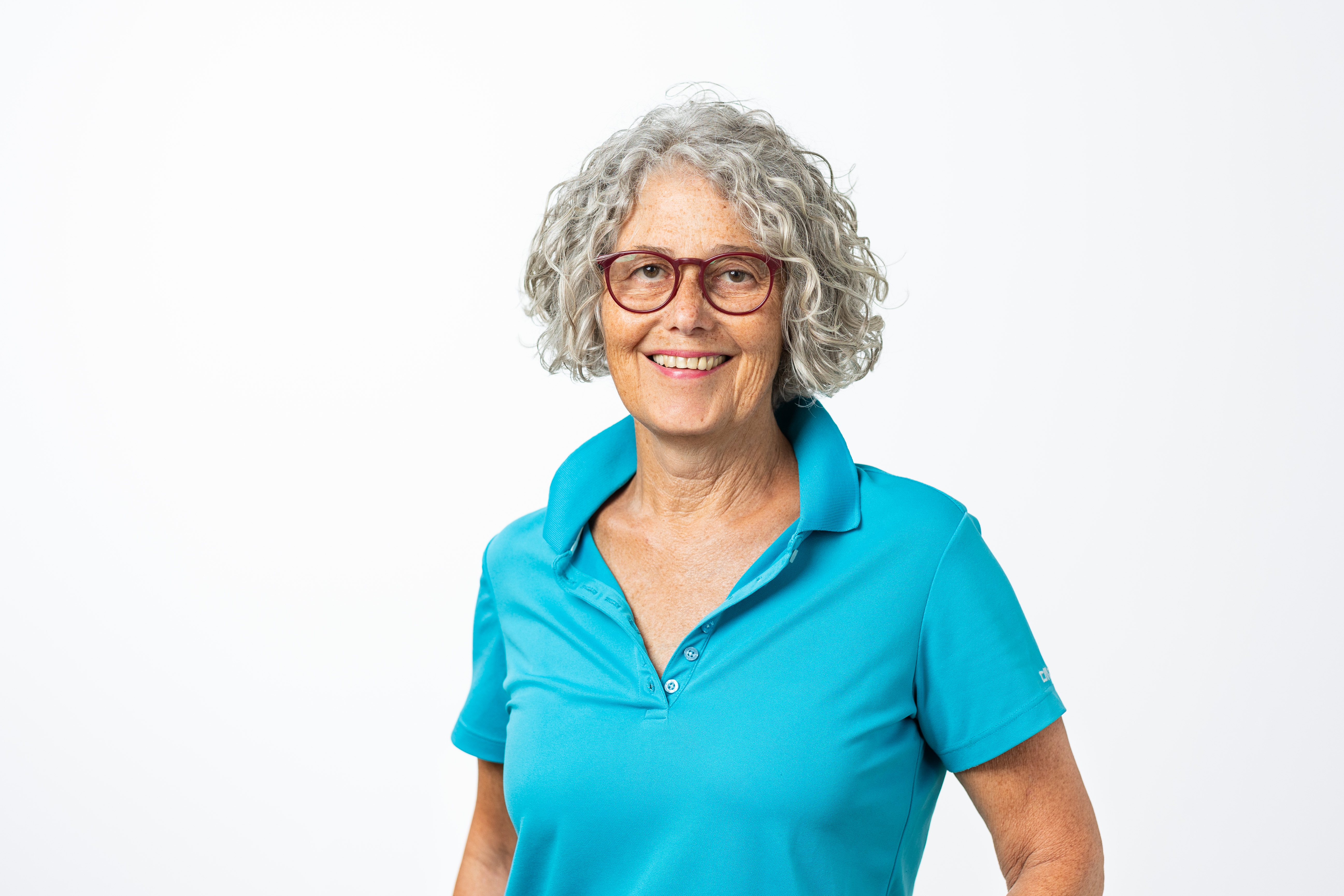 Margit Geiger trägt ein türkisfarbenes Polo-Shirt, eine Brille mit roter Fassung und die grauen Locken auf Kinnlänge offen.
