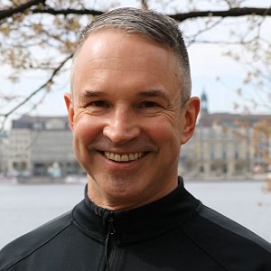 Porträtfoto des Journalisten und Autors Mike Powelz. Er steht im Freien, trägt eine schwarze Sportjacke und lacht in die Kamera.